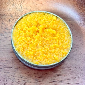 May include: A small round container filled with a bright yellow, grainy substance. The container is made of metal and has a silver lid.