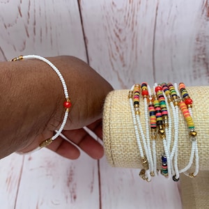 May include: A set of white beaded bracelets with gold and red accents. The bracelets are stacked on a beige fabric roll.