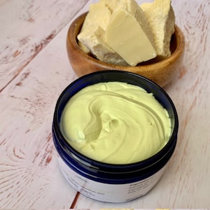May include: A close-up of a jar of yellow body butter with a blue lid. The jar is sitting on a wooden surface. There is a bowl of raw shea butter in the background.