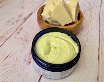 May include: A close-up of a jar of yellow body butter with a blue lid. The jar is sitting on a wooden surface. There is a bowl of raw shea butter in the background.