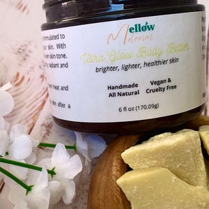 May include: A brown jar of Mellow Melanin Ultra Glow Body Butter. The jar is labeled with the product name, ingredients, and size: 6 fl oz (170.09g). The jar is sitting on a wooden surface with pieces of shea butter.