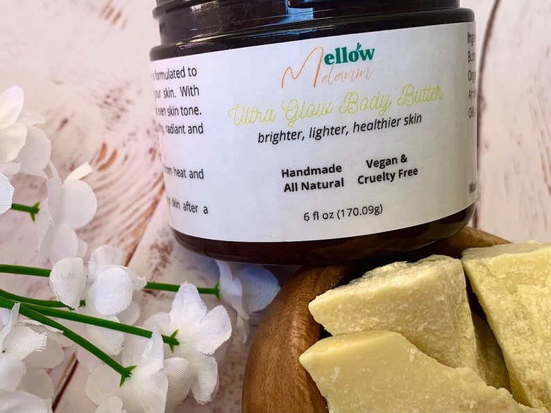 May include: A brown jar of Mellow Melanin Ultra Glow Body Butter. The jar is labeled with the product name, ingredients, and size: 6 fl oz (170.09g). The jar is sitting on a wooden surface with pieces of shea butter.