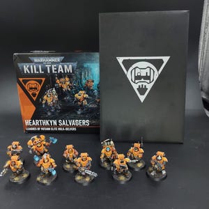 Puede incluir: Un set de caja Warhammer 40,000 Kill Team Hearthkyn Salvagers con Leagues of Votann elite hulk-delvers, acompañado de una caja de almacenamiento negra con un logotipo triangular blanco y diez figuras en miniatura pintadas con armadura naranja y gris.