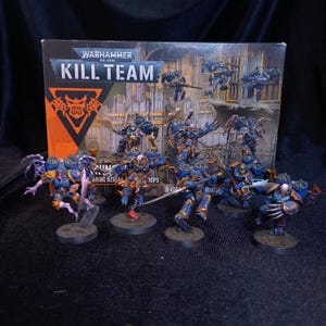 Op de afbeelding: Een Warhammer 40,000 Kill Team doos met een groep beschilderde miniatuurfiguren. De figuren zijn blauw en goud gepantserde krijgers met diverse wapens en vleugels, staand op ronde voetstukken voor de verpakking van het spel.