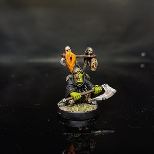 May include: A painted miniature figure of a green-skinned orc wearing a black hooded robe and holding a large silver axe. The figure features a wooden rack on its back adorned with two skulls and a long orange beard, all mounted on a round black base.