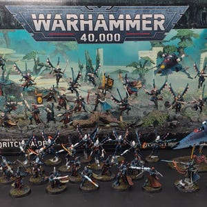Puede incluir: Una colección de figuras en miniatura de Warhammer 40,000 Aeldari Corsair pintadas, dispuestas frente a su caja original. El conjunto incluye varias unidades de infantería con accesorios alados y un modelo de avión azul sobre un soporte.