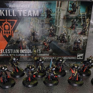 Puede incluir: Un set de caja Warhammer 40,000 Kill Team Celestian Insidiants con cazadores de brujas fanáticos de la Adepta Sororitas, acompañado de diez figuras en miniatura pintadas con armadura negra y roja con detalles dorados, sobre bases circulares.