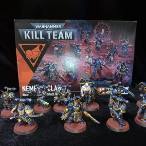 Puede incluir: Un set de caja Warhammer 40,000 Kill Team Nemesis Claw con un grupo de figuras en miniatura de space marines pintadas. Las figuras son principalmente azules y doradas con varios detalles de armadura y armas, sobre bases circulares frente a la caja.