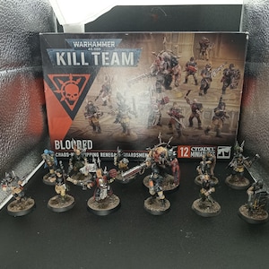 Puede incluir: Un set de caja Warhammer 40,000 Kill Team Blooded con guardias renegados adoradores del Caos, acompañado de doce figuras en miniatura pintadas sobre bases circulares, expuestas frente al embalaje original del producto.