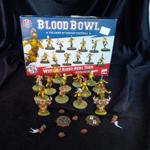 Puede incluir: Una caja de juego Blood Bowl Wood Elf Blood Bowl Team con los Athelorn Avengers. El set incluye una colección de figuras miniatura de elfos pintadas con uniformes amarillos y verdes, junto con pequeñas fichas de juego y balones.
