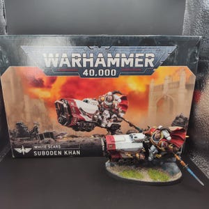 Puede incluir: Un set de miniaturas Warhammer 40,000 White Scars Suboden Khan que incluye una figura pintada sobre una motocicleta blanca y roja con una lanza, expuesta frente a su caja de cartón original con ilustraciones y texto de marca.
