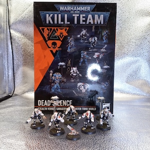 Puede incluir: Una caja de juego Warhammer 40,000 Kill Team Dead Silence con gráficos en naranja y negro y texto, acompañada de siete figuras en miniatura pintadas con armadura blanca, negra y roja sobre bases circulares frente a la caja.