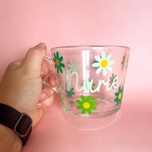 Puede incluir: Taza de vidrio transparente decorada con flores de margarita verdes y blancas y el nombre "Marie" en cursiva. La taza tiene un asa y se sostiene sobre un fondo rosa.