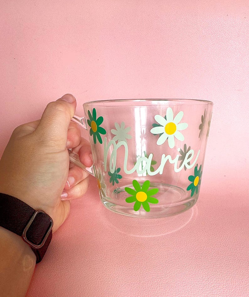 Puede incluir: Taza de vidrio transparente decorada con flores de margarita verdes y blancas y el nombre "Marie" en cursiva. La taza tiene un asa y se sostiene sobre un fondo rosa.
