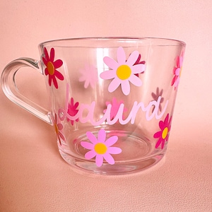 Puede incluir: Taza de vidrio transparente decorada con flores de margarita rosas y el nombre "Laura" en escritura rosa. Las flores tienen centros amarillos. La taza tiene un asa y est&aacute; sobre una superficie rosa.