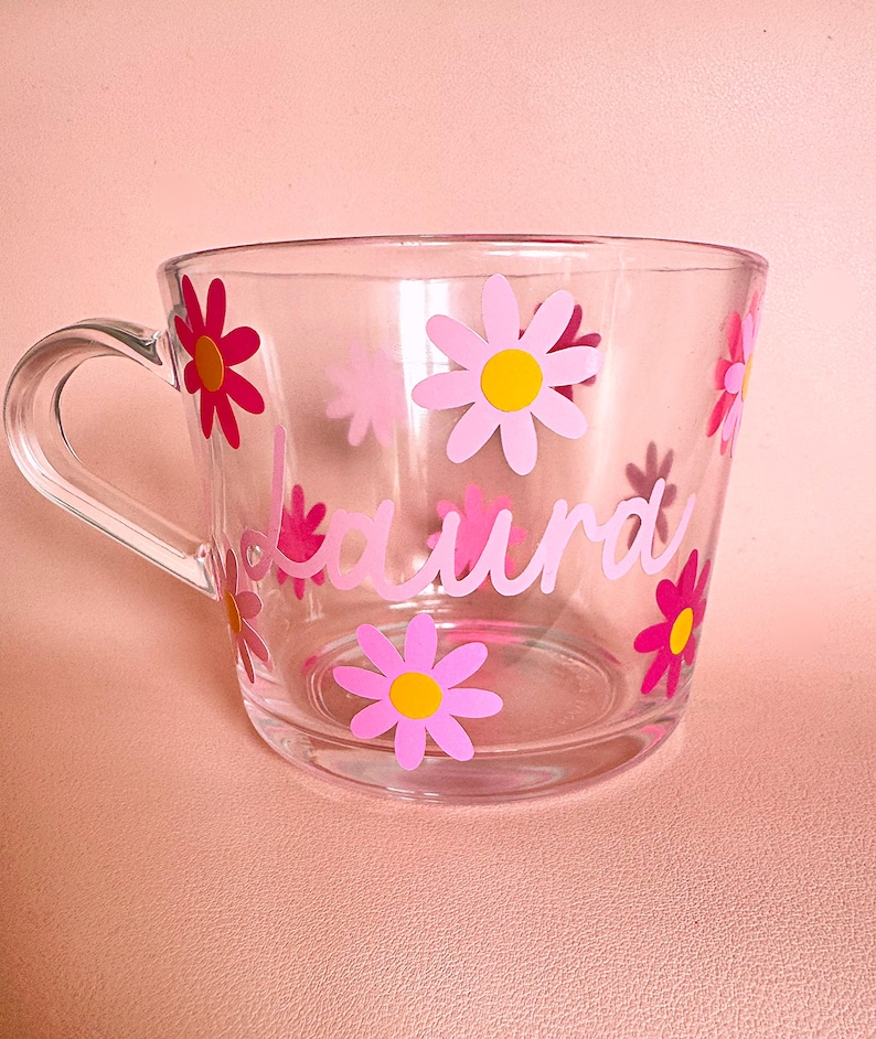 Puede incluir: Taza de vidrio transparente decorada con flores de margarita rosas y el nombre "Laura" en escritura rosa. Las flores tienen centros amarillos. La taza tiene un asa y est&aacute; sobre una superficie rosa.