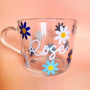 Puede incluir: Taza de vidrio transparente decorada con calcoman&iacute;as de flores de margarita de colores. La palabra "Rose" est&aacute; escrita en cursiva en el lateral. La taza tiene un asa y est&aacute; sobre un fondo color melocot&oacute;n.