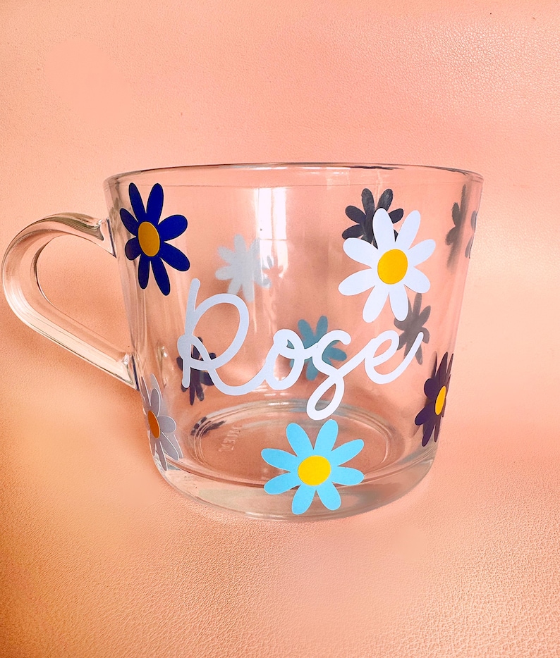 Puede incluir: Taza de vidrio transparente decorada con calcoman&iacute;as de flores de margarita de colores. La palabra "Rose" est&aacute; escrita en cursiva en el lateral. La taza tiene un asa y est&aacute; sobre un fondo color melocot&oacute;n.