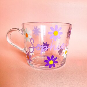 Puede incluir: Taza de cristal transparente decorada con flores de margarita moradas, blancas y amarillas. El nombre "Sophie" est&aacute; escrito en p&uacute;rpura en la parte delantera de la taza. La taza tiene un asa transparente y est&aacute; sobre un fondo rosa.