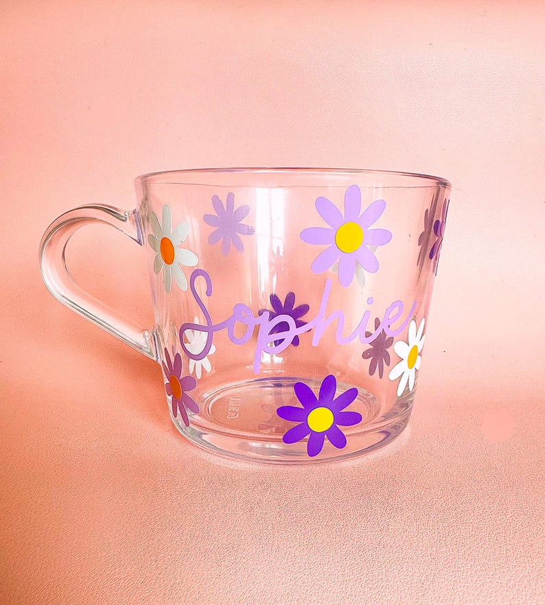 Puede incluir: Taza de cristal transparente decorada con flores de margarita moradas, blancas y amarillas. El nombre "Sophie" est&aacute; escrito en p&uacute;rpura en la parte delantera de la taza. La taza tiene un asa transparente y est&aacute; sobre un fondo rosa.