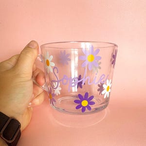 Puede incluir: Taza de vidrio transparente decorada con calcoman&iacute;as de flores de margarita moradas, amarillas y blancas. El nombre "Sophie" est&aacute; escrito en p&uacute;rpura en el lateral de la taza. La taza tiene un asa y est&aacute; sobre un fondo rosa.