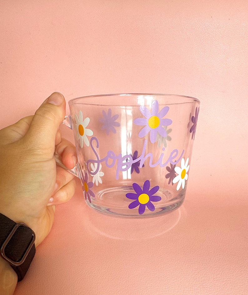 Puede incluir: Taza de vidrio transparente decorada con calcoman&iacute;as de flores de margarita moradas, amarillas y blancas. El nombre "Sophie" est&aacute; escrito en p&uacute;rpura en el lateral de la taza. La taza tiene un asa y est&aacute; sobre un fondo rosa.