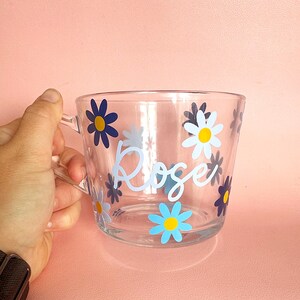 Puede incluir: Taza de vidrio transparente con asa, decorada con calcoman&iacute;as de flores de margarita azules, amarillas y blancas. El nombre "Rose" est&aacute; escrito en cursiva en la parte delantera. La taza est&aacute; sobre un fondo rosa.