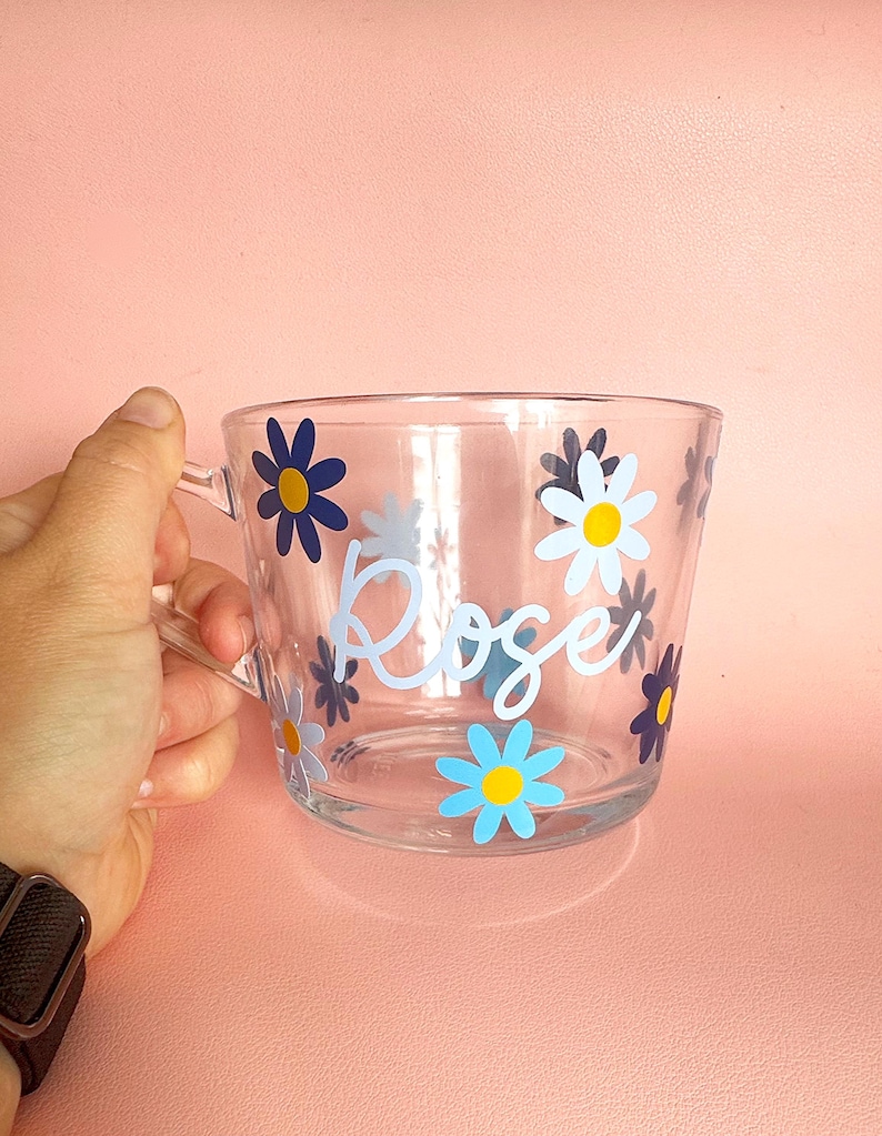 Puede incluir: Taza de vidrio transparente con asa, decorada con calcoman&iacute;as de flores de margarita azules, amarillas y blancas. El nombre "Rose" est&aacute; escrito en cursiva en la parte delantera. La taza est&aacute; sobre un fondo rosa.