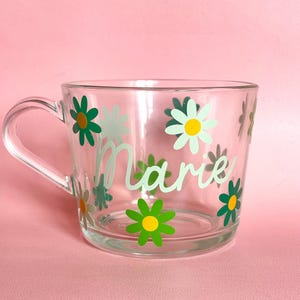 Puede incluir: Taza de vidrio transparente con asa, decorada con dise&ntilde;os de flores de margarita verdes y blancas. El nombre "Marie" est&aacute; escrito en una escritura verde claro en la parte delantera. La taza est&aacute; sobre un fondo rosa.