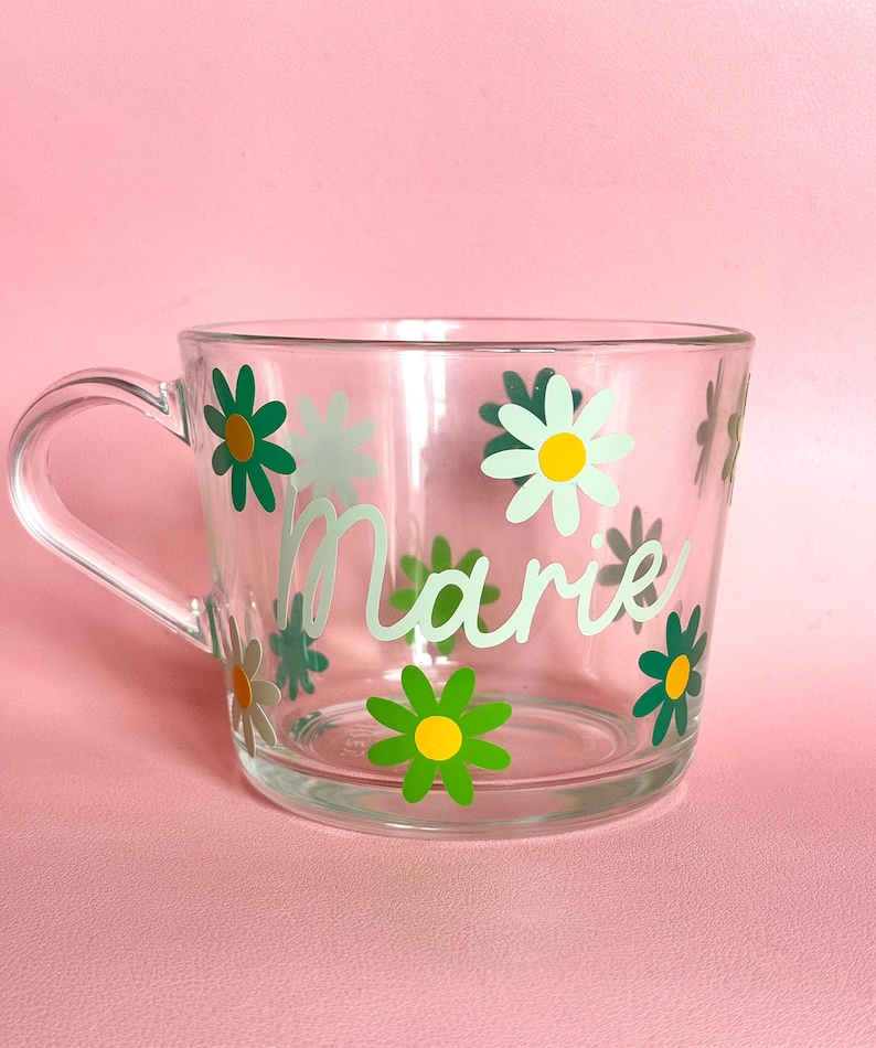 Puede incluir: Taza de vidrio transparente con asa, decorada con dise&ntilde;os de flores de margarita verdes y blancas. El nombre "Marie" est&aacute; escrito en una escritura verde claro en la parte delantera. La taza est&aacute; sobre un fondo rosa.
