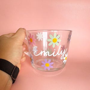 Puede incluir: Taza de vidrio transparente decorada con flores de margarita coloridas y el nombre "Emily" en escritura blanca. La taza tiene un asa y se sostiene sobre un fondo rosa.