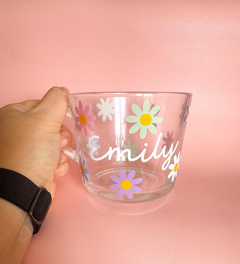 Puede incluir: Taza de vidrio transparente decorada con flores de margarita coloridas y el nombre "Emily" en escritura blanca. La taza tiene un asa y se sostiene sobre un fondo rosa.