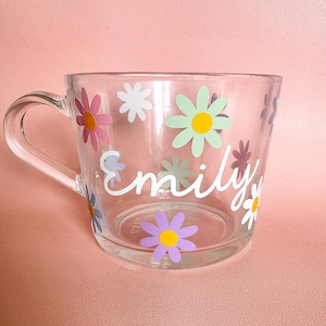 Puede incluir: Taza de vidrio transparente con asa, decorada con flores de margarita coloridas y el nombre "Emily" en escritura blanca. Las margaritas son en tonos rosa, verde, morado y blanco, con centros amarillos. La taza est&aacute; sobre una superficie rosa.
