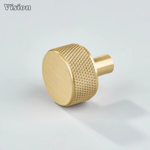 May include: A gold-colored cabinet knob with a textured, diamond-patterned grip. The knob has a cylindrical shape with a flat, circular face and a short, cylindrical stem. The word "Vision" is in the upper left corner.