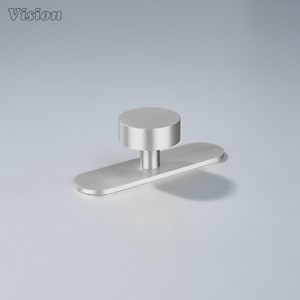 Op de afbeelding: Een geborstelde zilveren kastknop met een cilindrische bovenkant en een rechthoekige basis. De knop is gemonteerd op een lichtgrijze ondergrond. Het woord "Vision" staat in de linkerbovenhoek.