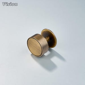May include: A bronze-colored, cylindrical curtain rod finial with a circular base and a flat, round top. The finial has a vintage aesthetic and is designed for decorative purposes. The word "Vision" is printed in the top left corner.