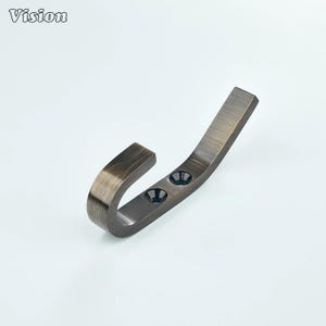 Pode incluir: Um gancho de metal de cor bronze com um design curvo. O gancho tem dois orifícios para parafusos e a palavra "Vision" no canto superior esquerdo. O gancho foi projetado para pendurar itens.