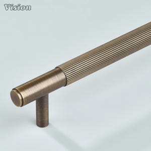May include: Bronze-toned cabinet pull with a ribbed handle and a cylindrical end. The pull has a T-shaped base for mounting. The word "Vision" is in the upper left corner. This hardware is suitable for kitchen or bathroom cabinets.