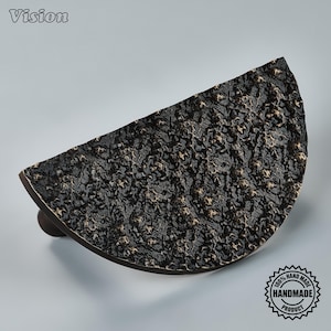 May include: A semi-circular cabinet pull with a dark, textured, rugged surface featuring bronze-colored highlights. The hardware has a distinct half-moon shape and a rough, uneven finish across its front face, providing a tactile appearance.