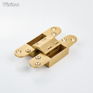 Pode incluir: Uma dobradiça oculta de metal em tom dourado escovado com um mecanismo de pivô central e quatro placas de montagem arredondadas, cada uma contendo dois orifícios para parafusos escareados. A palavra Vision está impressa no canto superior esquerdo.
