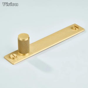 May include: A gold-toned cabinet pull with a textured cylindrical handle and a rectangular base. The base has two screws for mounting. The word "Vision" is in the upper left corner.