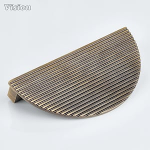 May include: A half-moon shaped cabinet pull with a bronze finish. The pull features a series of parallel, horizontal lines across its surface. The word "Vision" is in the upper left corner.