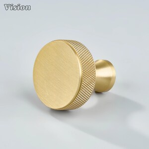 May include: A gold-colored cabinet knob with a circular design. The knob features a textured, diamond-patterned edge and a smooth, flat center. The Vision logo is in the top left corner.