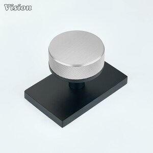 Pode incluir: Um puxador de móvel com um topo circular prateado com uma borda serrilhada texturizada, montado numa placa de base retangular preta fosca. A palavra Vision está impressa no canto superior esquerdo da imagem numa fonte preta simples.