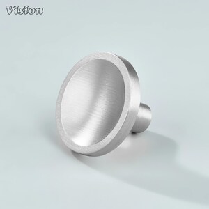 May include: A brushed metal cabinet knob with a circular, concave design. The knob has a cylindrical stem and a wide, flat face. The word "Vision" is in the upper left corner. The hardware is silver in colour.
