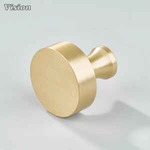 Puede incluir: Un pomo de armario de color dorado con una cara circular y un vástago cilíndrico. El pomo tiene un acabado de metal cepillado y está sobre un fondo claro. La palabra "Vision" está en la esquina superior izquierda.