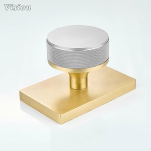 Pode incluir: Um puxador de móvel com um topo circular em tom prateado com uma borda serrilhada texturizada, montado sobre uma haste cilíndrica dourada e uma placa de base retangular dourada. A palavra Vision está impressa no canto superior esquerdo.