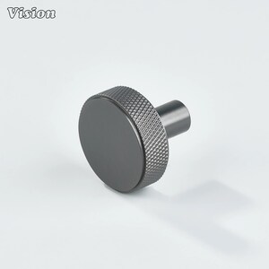May include: A dark gray cabinet knob with a circular face and a textured, diamond-patterned edge. The knob has a cylindrical stem and the word "Vision" is printed in the upper left corner. This hardware is suitable for drawers and cabinets.
