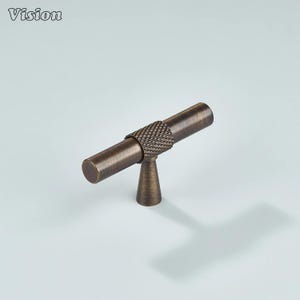May include: Bronze-toned cabinet pull with a T-bar design. The center section features a textured, diamond-patterned grip. The Vision logo is in the upper left corner. The pull is mounted on a light blue background.