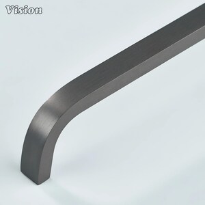 Pode incluir: Uma puxador de metal cinza escuro curvo com acabamento escovado. A palavra "Vision" está impressa no canto superior esquerdo. O puxador tem formato retangular e um design suave e moderno, adequado para armários ou gavetas.